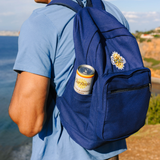 A navy blue canvas backpack worn by a person outdoors near the ocean, with a can of Surfside Lemonade in the side pocket and a Surfside logo patch on the back.