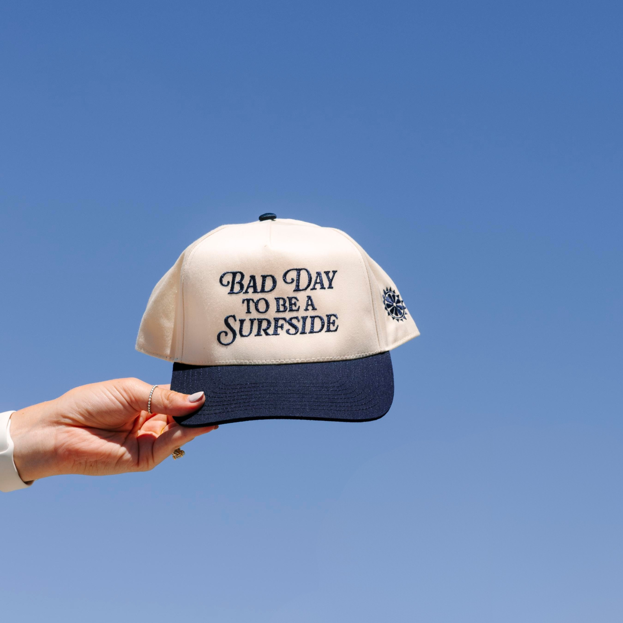 Cream and navy cap with 'Bad Day to be a Surfside' text held against a clear blue sky