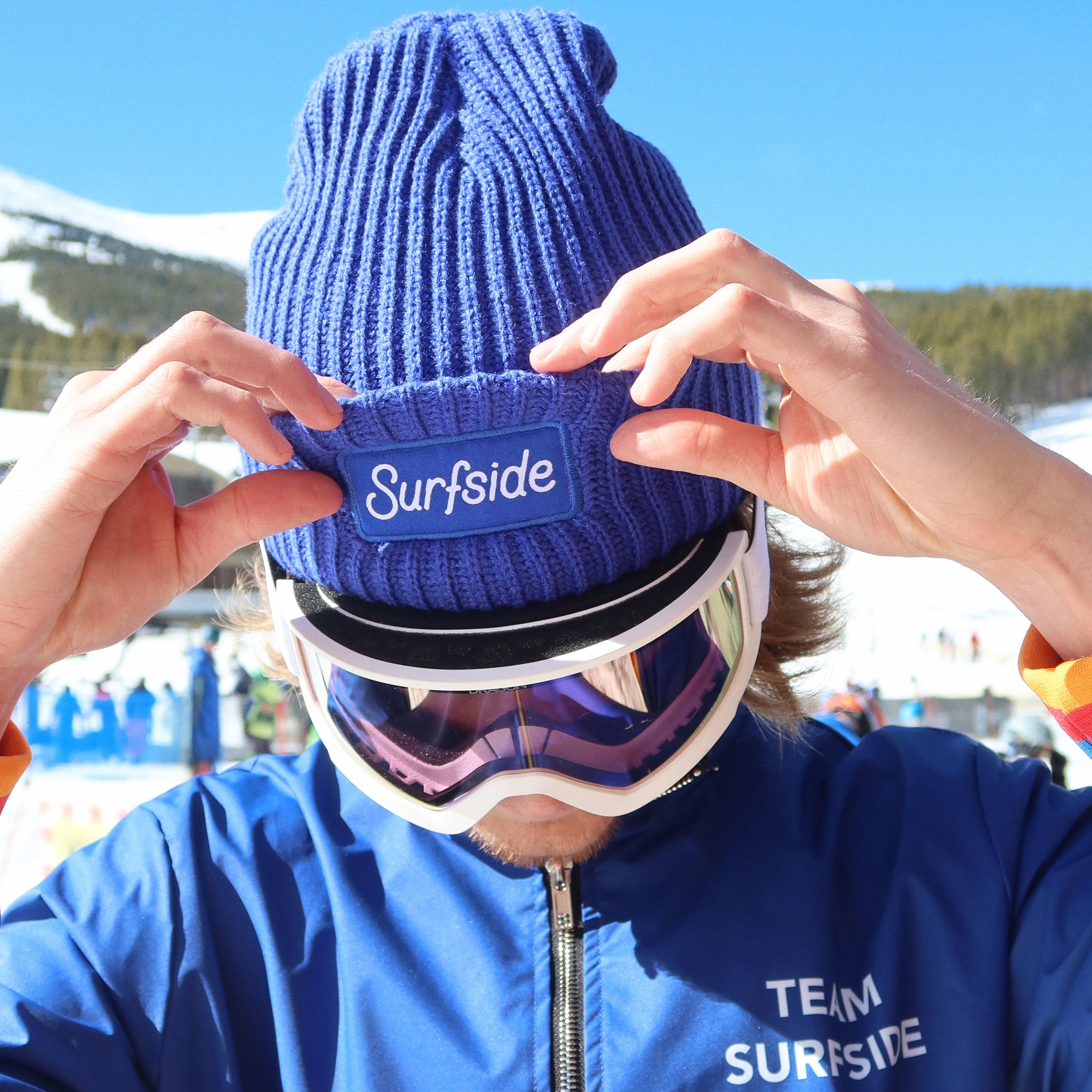 Person wearing a blue 'Surfside' beanie and goggles with a blue jacket, against a snowy background.