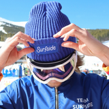 Person wearing a blue 'Surfside' beanie and goggles with a blue jacket, against a snowy background.