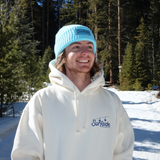 Person wearing a cream hoodie and light blue beanie standing in a snowy landscape with trees.