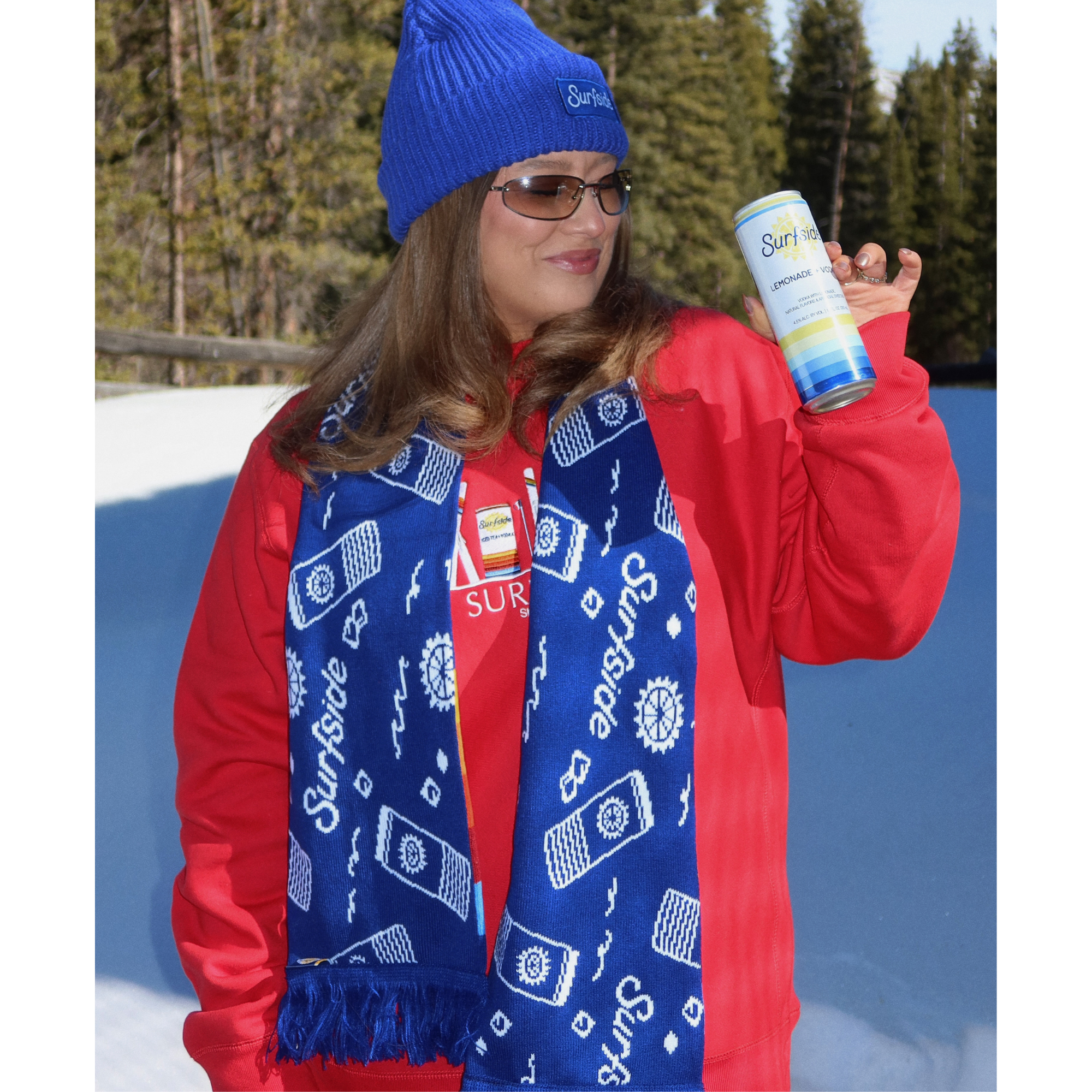 A woman in a red jacket, blue beanie, and blue patterned scarf holds up a can of Surfside Hot Chocolate outdoors in the snow with trees in the background.
