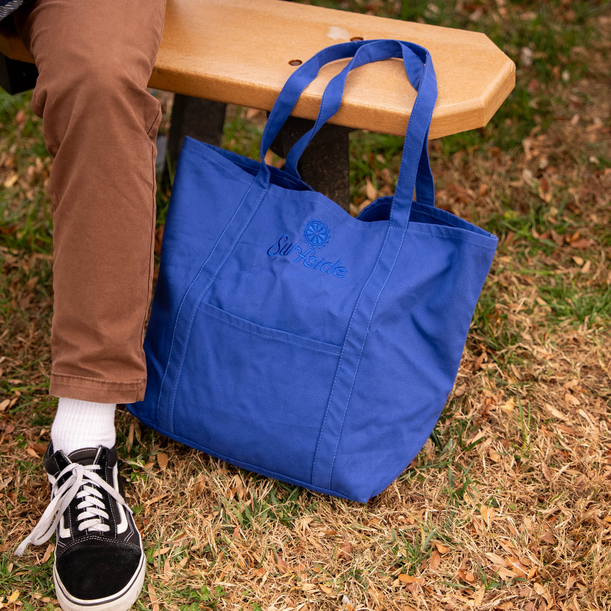 A close up of the cobalt blue embroidered Surfside tote bag laying in grass with someone sitting on a bench wearing sneakers