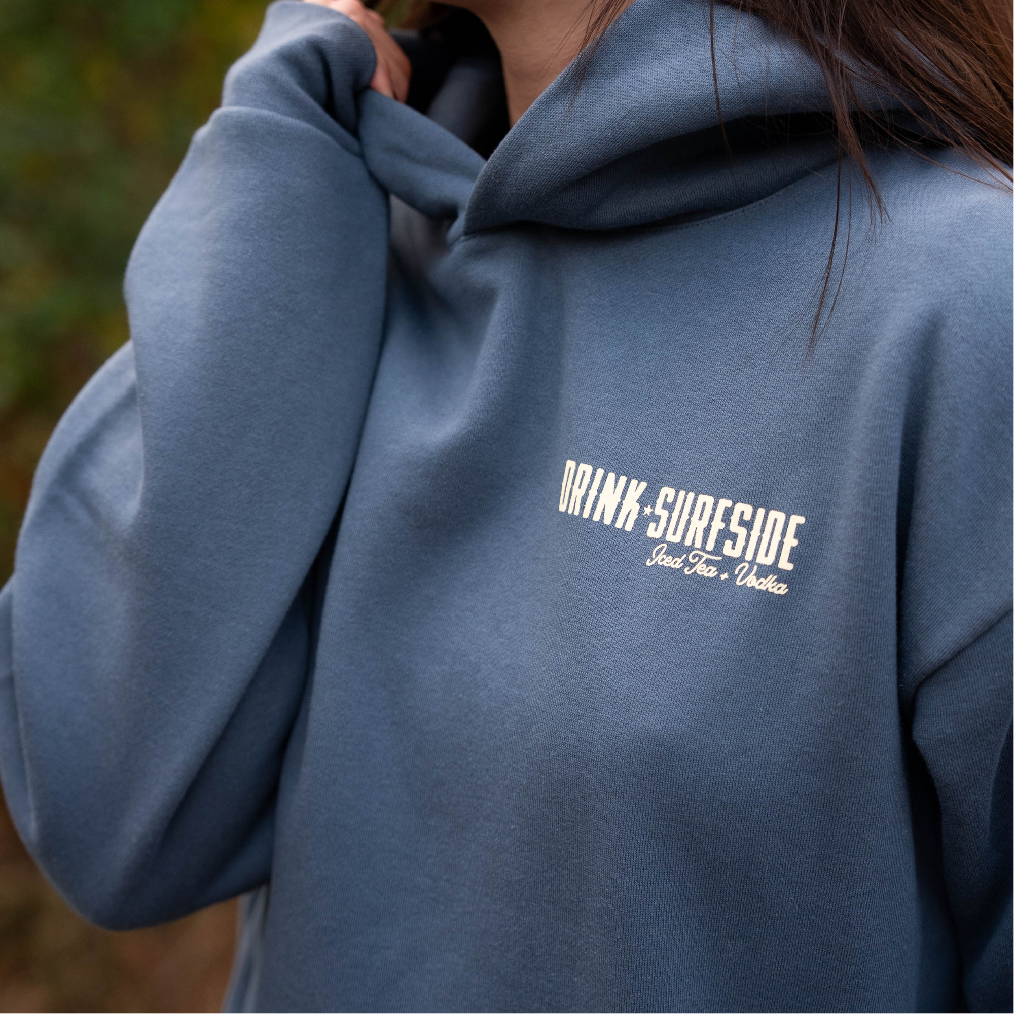 Person wearing a blue hoodie with western inspired Drink Surfside Ice Tea and Vodka text, standing outdoors.