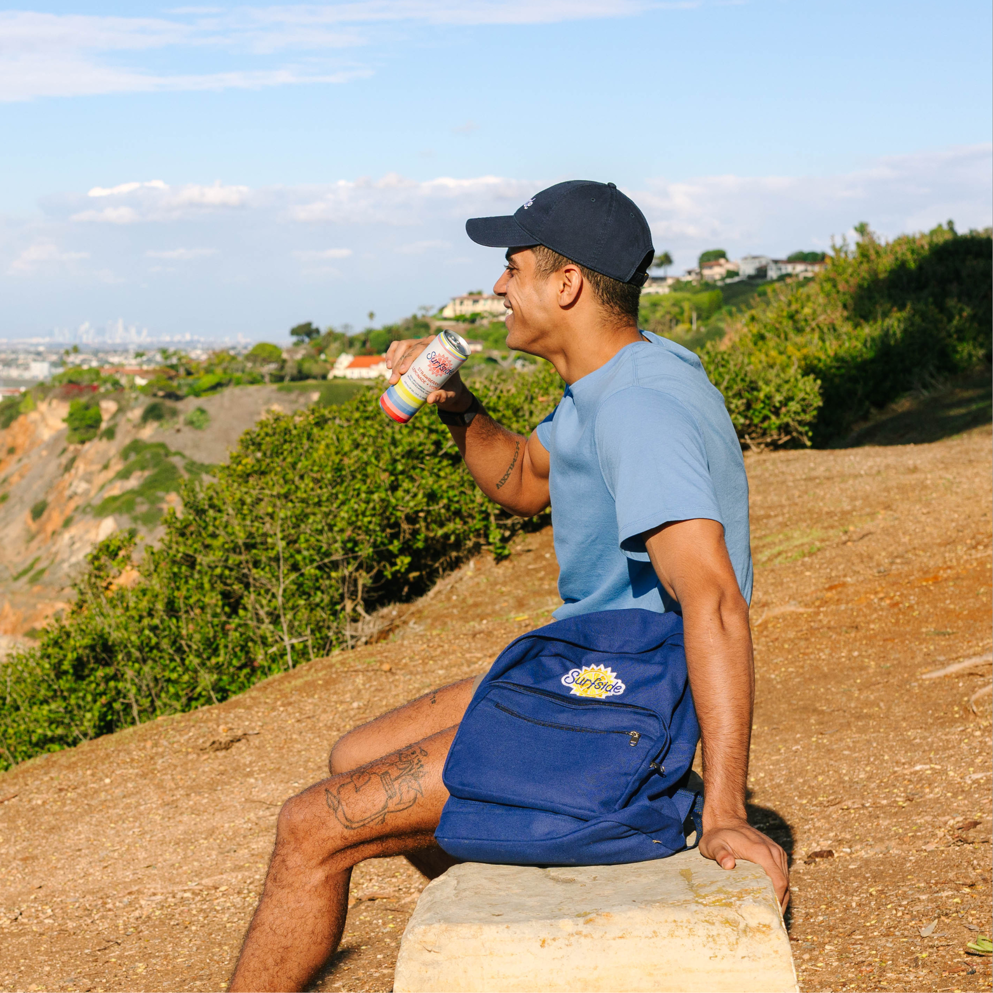 A man sitting on a stone bench outdoors on a hillside, wearing a light blue t-shirt, navy blue shorts with a yellow and white logo, and a navy blue baseball cap. He is holding a can of Surfside.