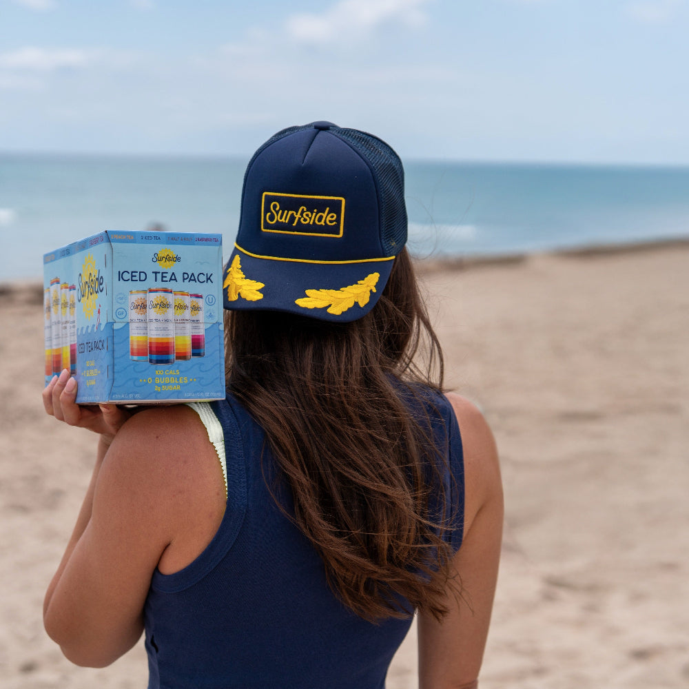 Person wearing a navy blue captains hat with yellow rope around the brim and yellow detailing holding a variety pack of Surfside Iced Teas in an outdoor beach setting