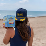 Person wearing a navy blue captains hat with yellow rope around the brim and yellow detailing holding a variety pack of Surfside Iced Teas in an outdoor beach setting