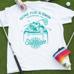 White t-shirt with 'Gone for a Swim' graphic on grass with golf equipment