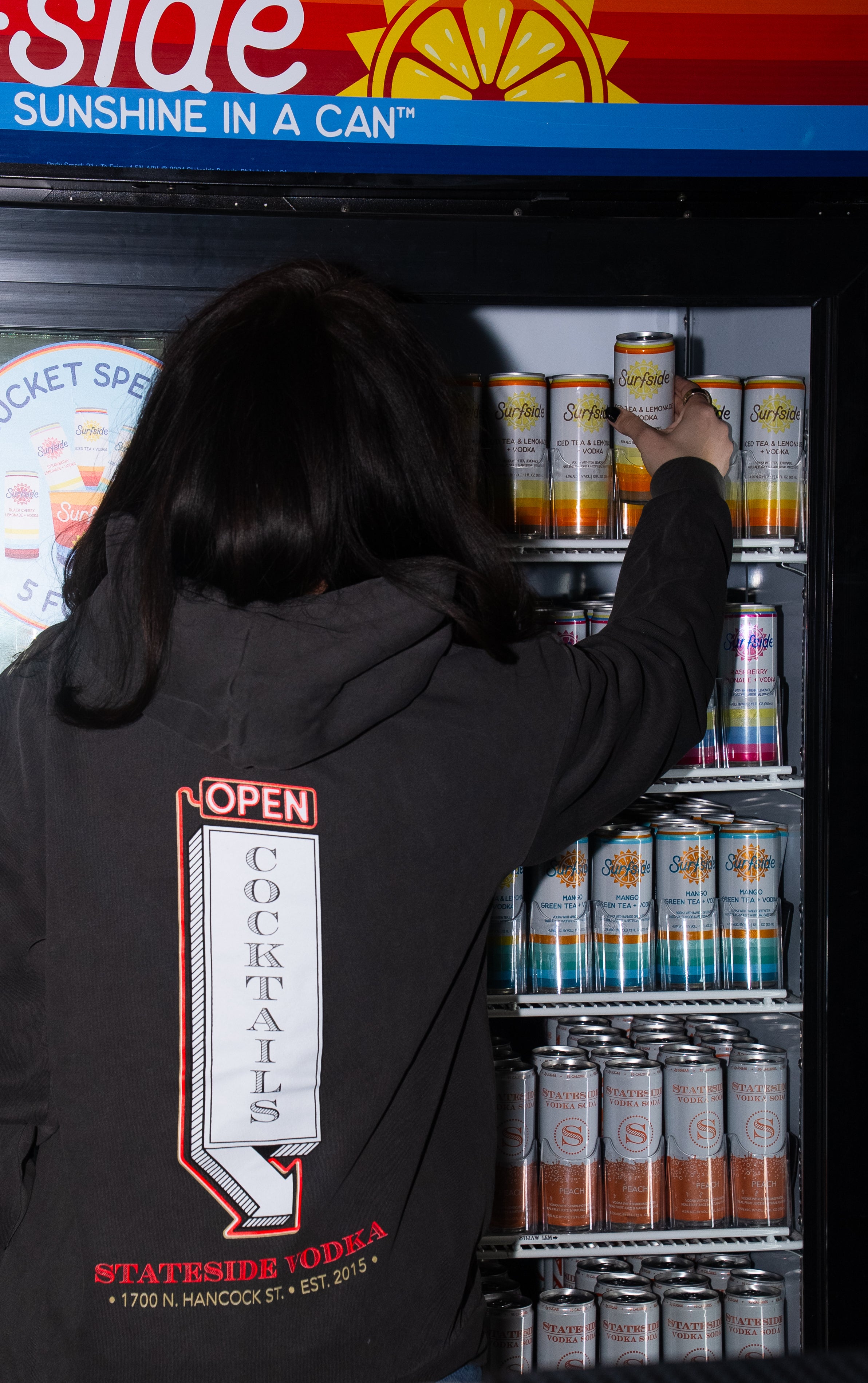 Person reaching into a refrigerator  filled with Surfside cans, with a 'Sunshine in a Can' sign above.