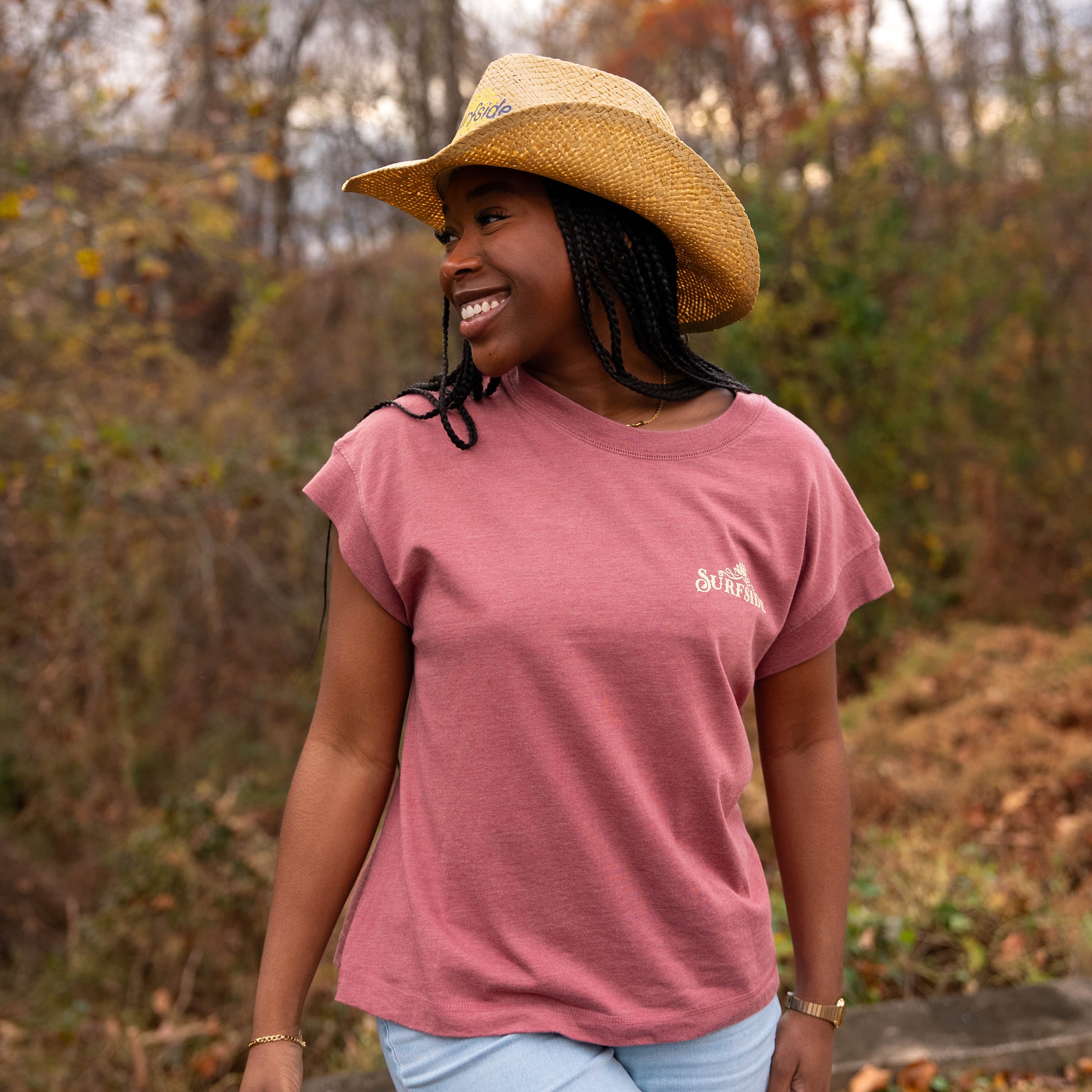 Woman wearing a faded red t-shirt with a western inspired Surfside logo and straw hat in an outdoor setting with trees and foliage.