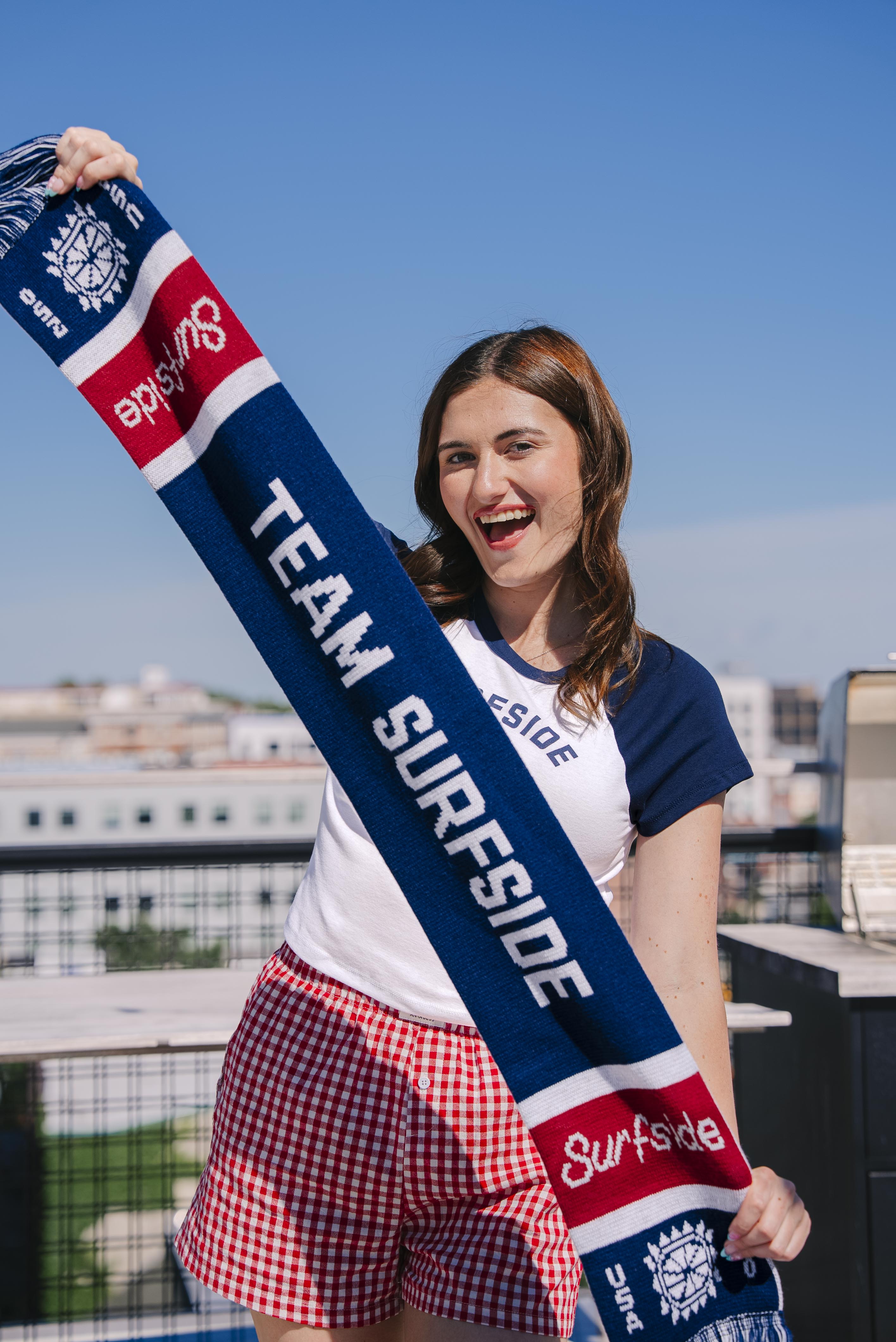 Girl holding Team Surfside USA 250 themed scarf. The Soccer scarf features Team Surfside in the middle, the surfside logo and the sun logo with fringe. The soccer scarf is red, white and blue.