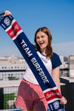 Girl holding Team Surfside USA 250 themed scarf. The Soccer scarf features Team Surfside in the middle, the surfside logo and the sun logo with fringe. The soccer scarf is red, white and blue.