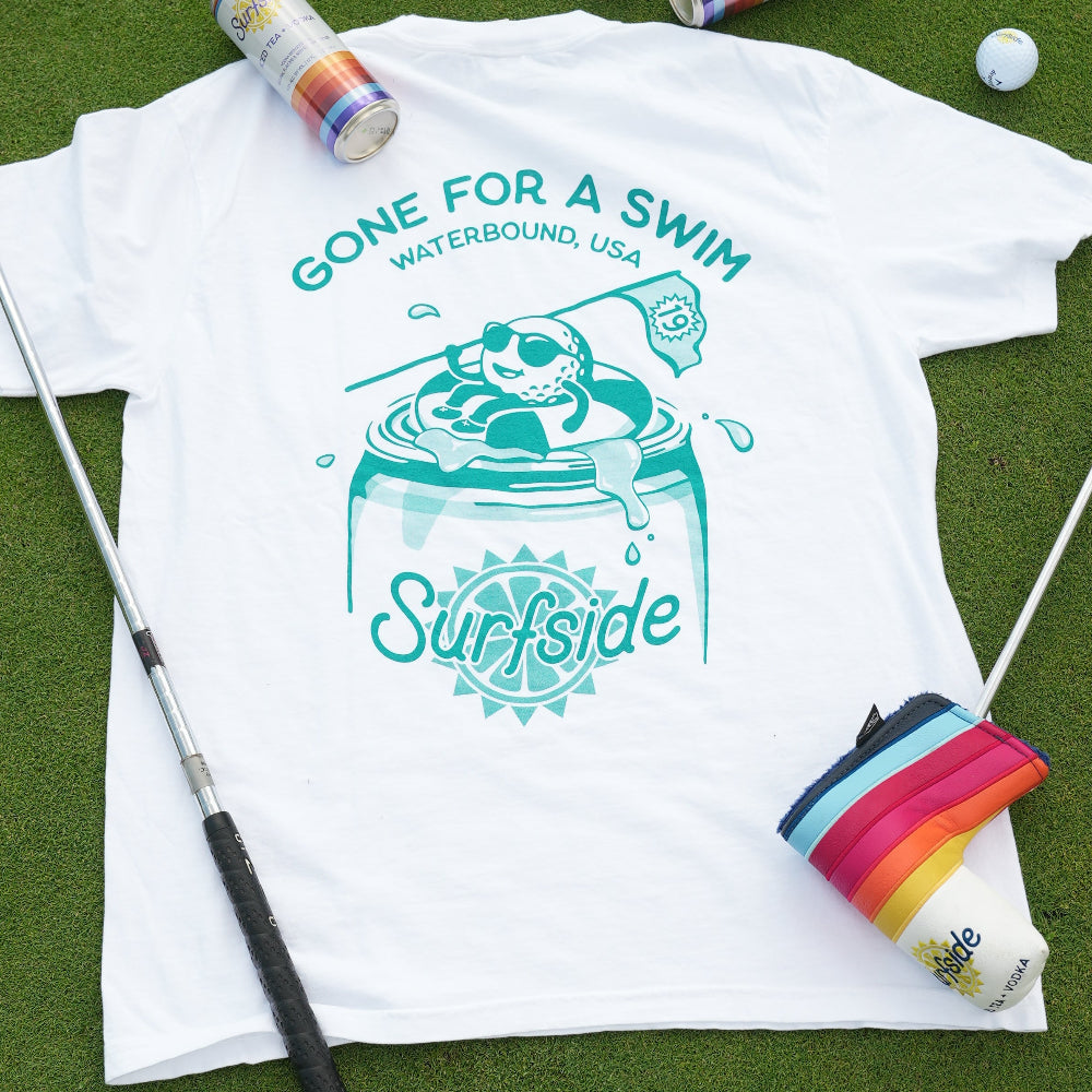 White t-shirt with '19th Hole Gone for a Swim Waterbound, USA Surfside' graphic on grass turf surrounded by golf equipment