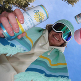 Man holding Surfside in a green Surfside patched beanie and a green scarf looking down in the camera with a light blue sky in the background.