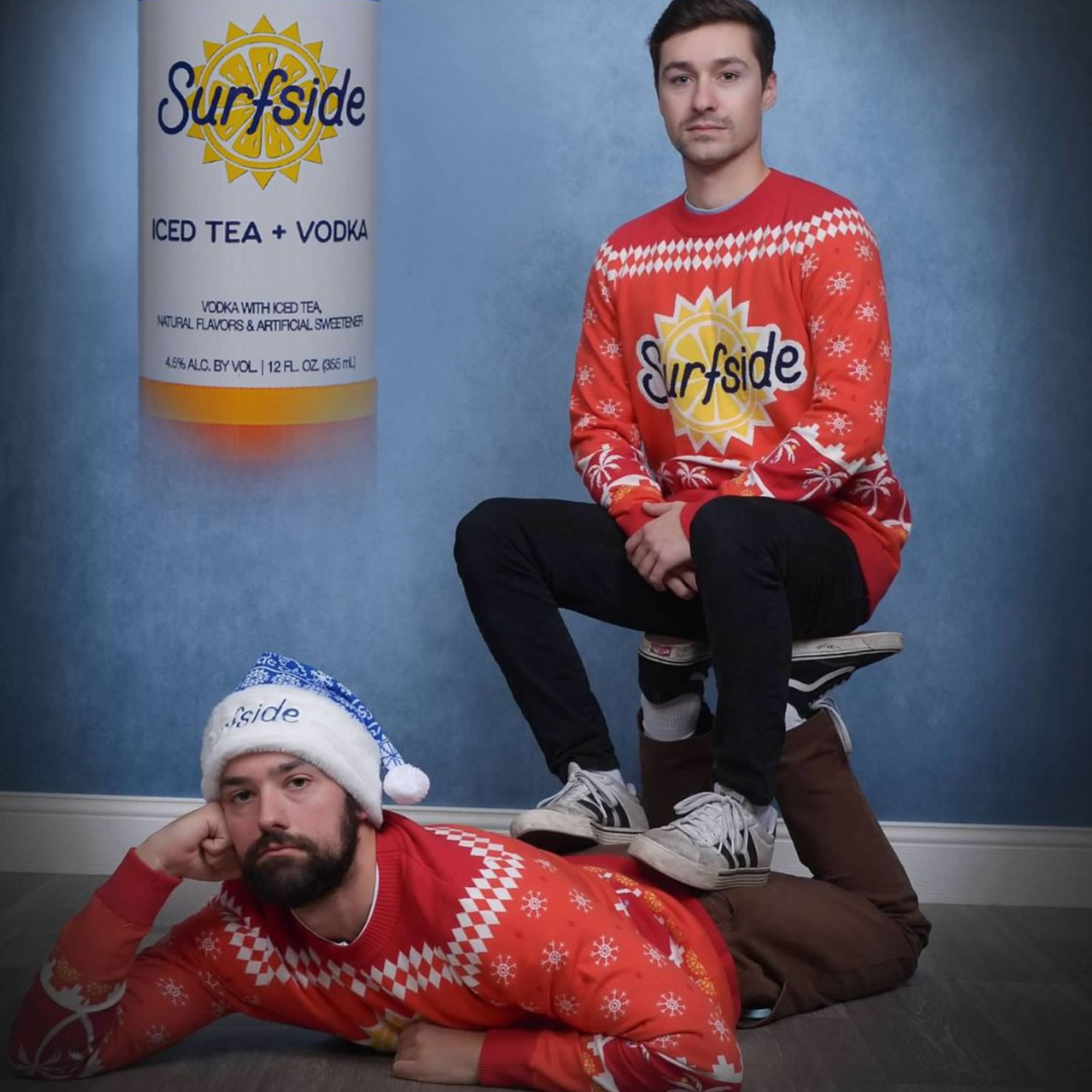 Two men posing in red and white holiday sweaters featuring the 'Surfside' logo, set against a blue textured wall with a large 'Surfside' can graphic on the left.