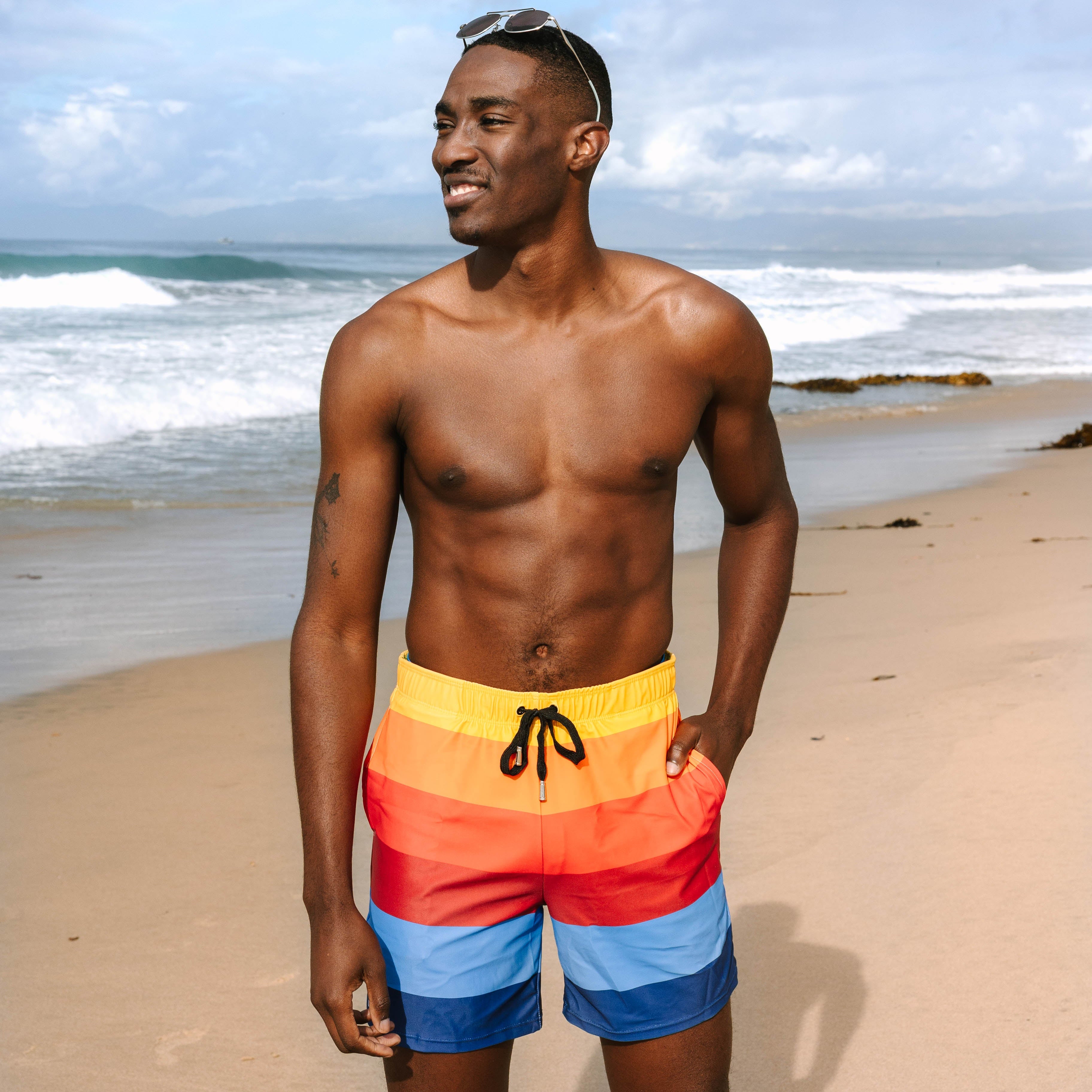 Man standing on a beach wearing Surfside branded swim trunks with ocean waves in the background.