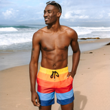 A man standing on a sandy beach wearing colorful horizontal-striped swim trunks with yellow, orange, red, light blue, and dark blue bands, with a black drawstring.