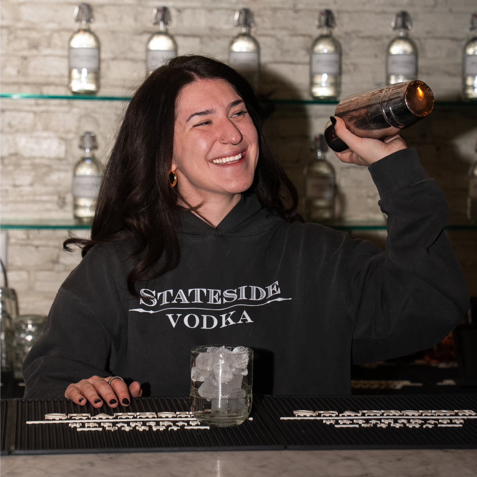 A woman smiling and holding a metal cocktail shaker behind a bar, wearing a black hoodie with 'STATESIDE VODKA' printed in white across the chest. A glass with ice sits on the bar, and shelves with bottles are visible in the background.