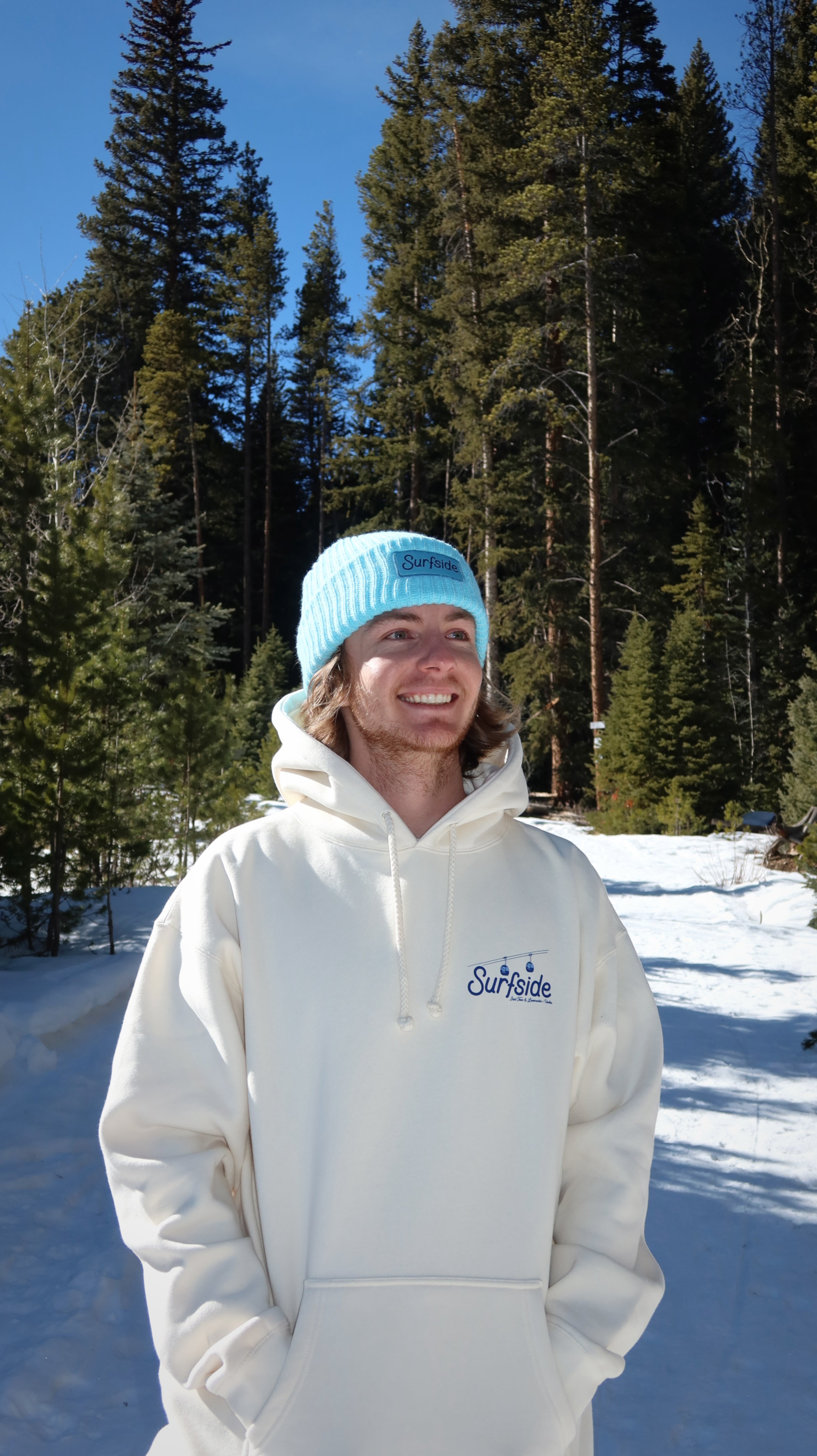 Person wearing a cream hoodie and light blue beanie standing in a snowy landscape with trees.