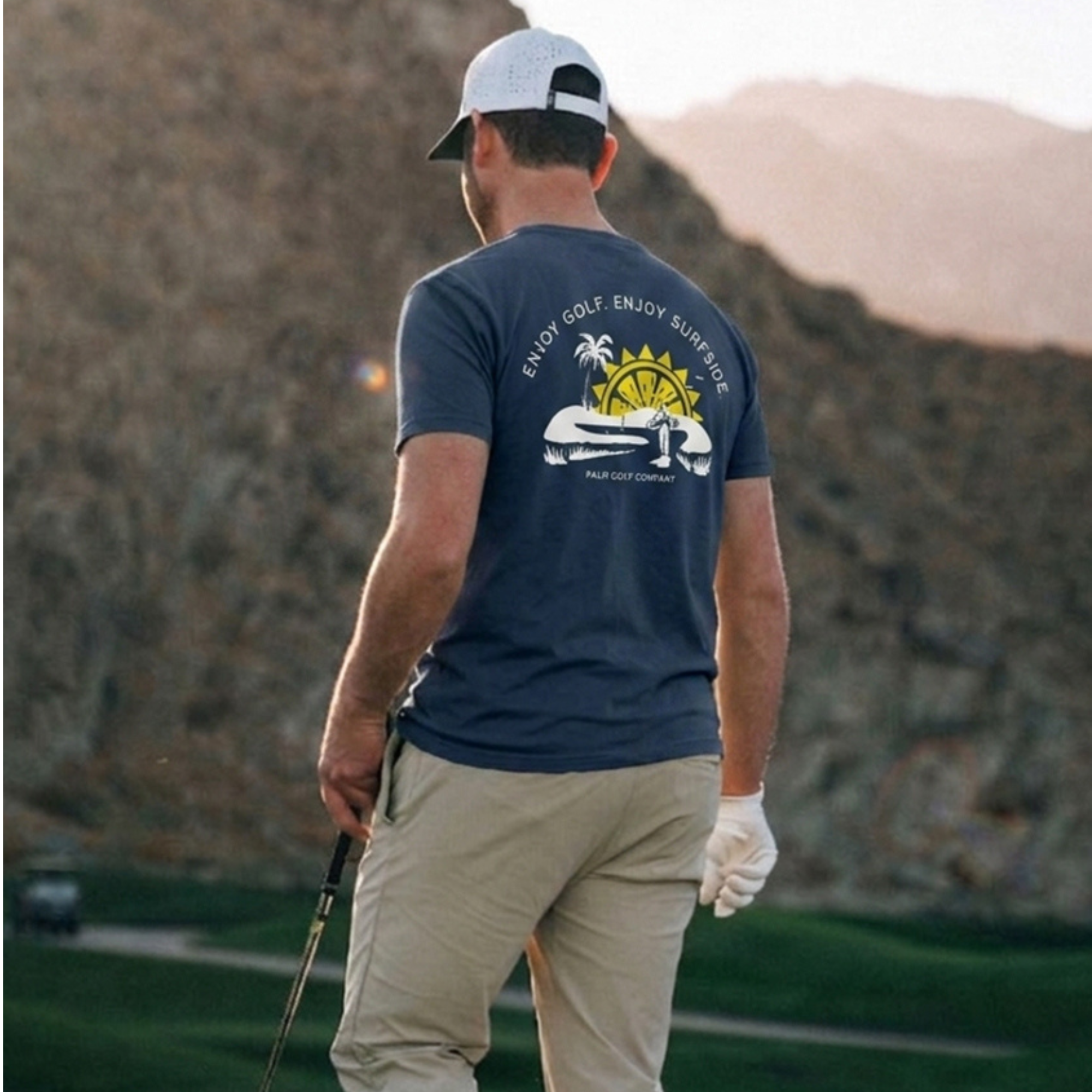 Back view of a man in a scenic background wearing a navy blue Surfside and Palm Golf T-shirt design featuring a golf course, sun, palm tree and 