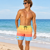 A man wearing colorful horizontal-striped swim trunks walks on a sandy beach with the ocean in the background. The trunks have stripes of yellow, orange, coral, and light blue, with a lime green waistband and a small yellow logo on the left leg.