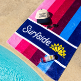 A striped beach towel laid poolside, featuring the Surfside logo and a yellow sun graphic. A cream baseball cap with blue text featuring "BAD DAY TO BE A SURFSIDE", sunglasses, and a Surfside Raspberry Tea + Vodka can that are placed on top of the towel