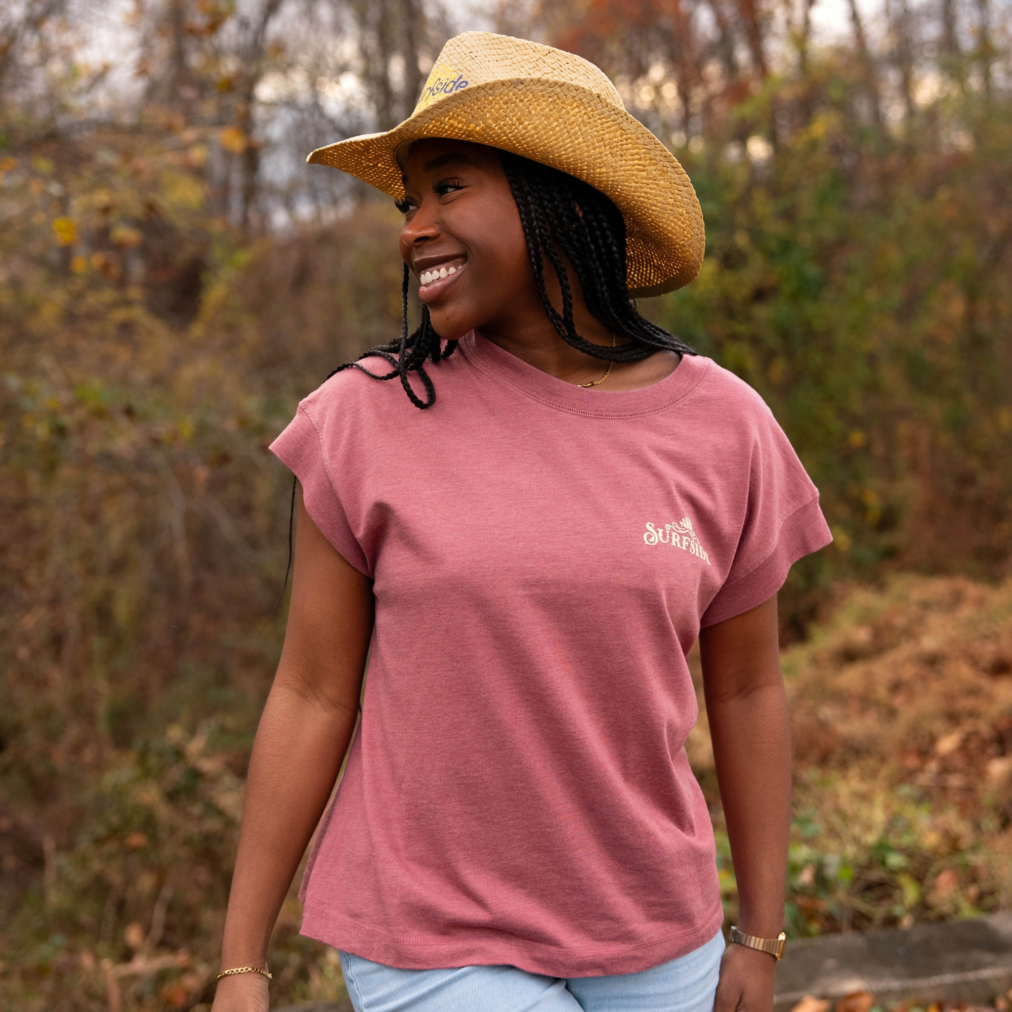 Woman wearing a faded red t-shirt with a western inspired Surfside logo and straw hat in an outdoor setting with trees and foliage.