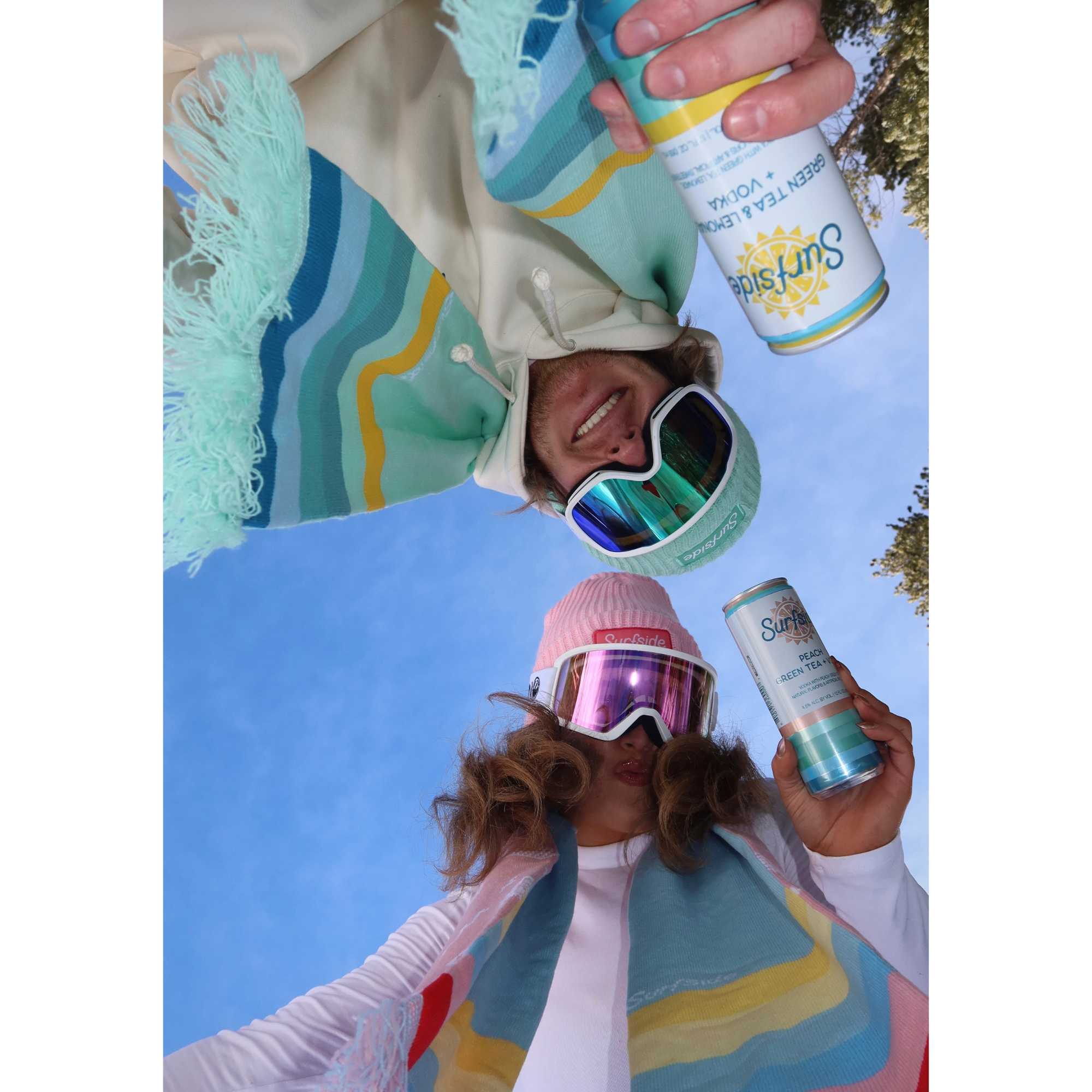Two people holding Surfside cans looking down with the Surfside colorful scarves around their neck and in the background there's a blue sky