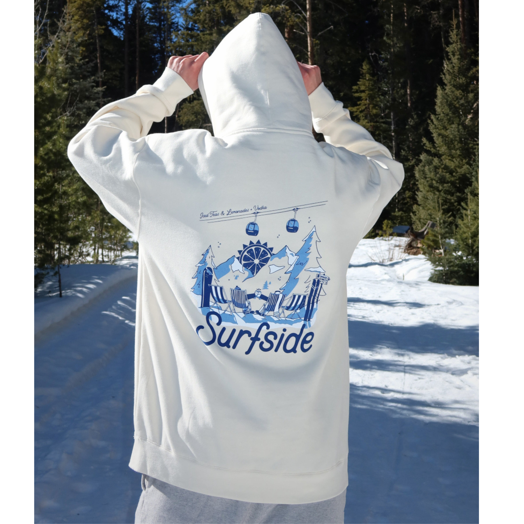A person standing in a snowy outdoor setting wearing a white hoodie, viewed from behind. The back of the hoodie has a large blue graphic print featuring mountains, surfboards, a snowboarder, and the word 'Surfside'.