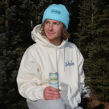 Person wearing a white hoodie and blue beanie sitting on a log with trees in the background