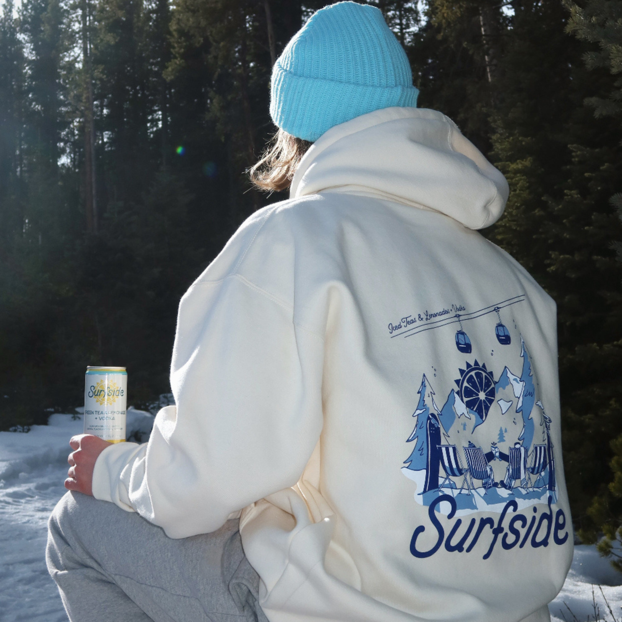 Person wearing a cream hoodie with a blue 'Surfside' design, sitting in a snowy landscape.