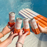 Four cans of Stateside Vodka Soda Orange being held by hands by a pool with Stateside Vodka Orange pool float in the background