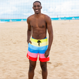 A man standing on a sandy beach wearing colorful horizontal-striped swim trunks with yellow, light blue, dark blue, pink, and red bands, with a black drawstring.