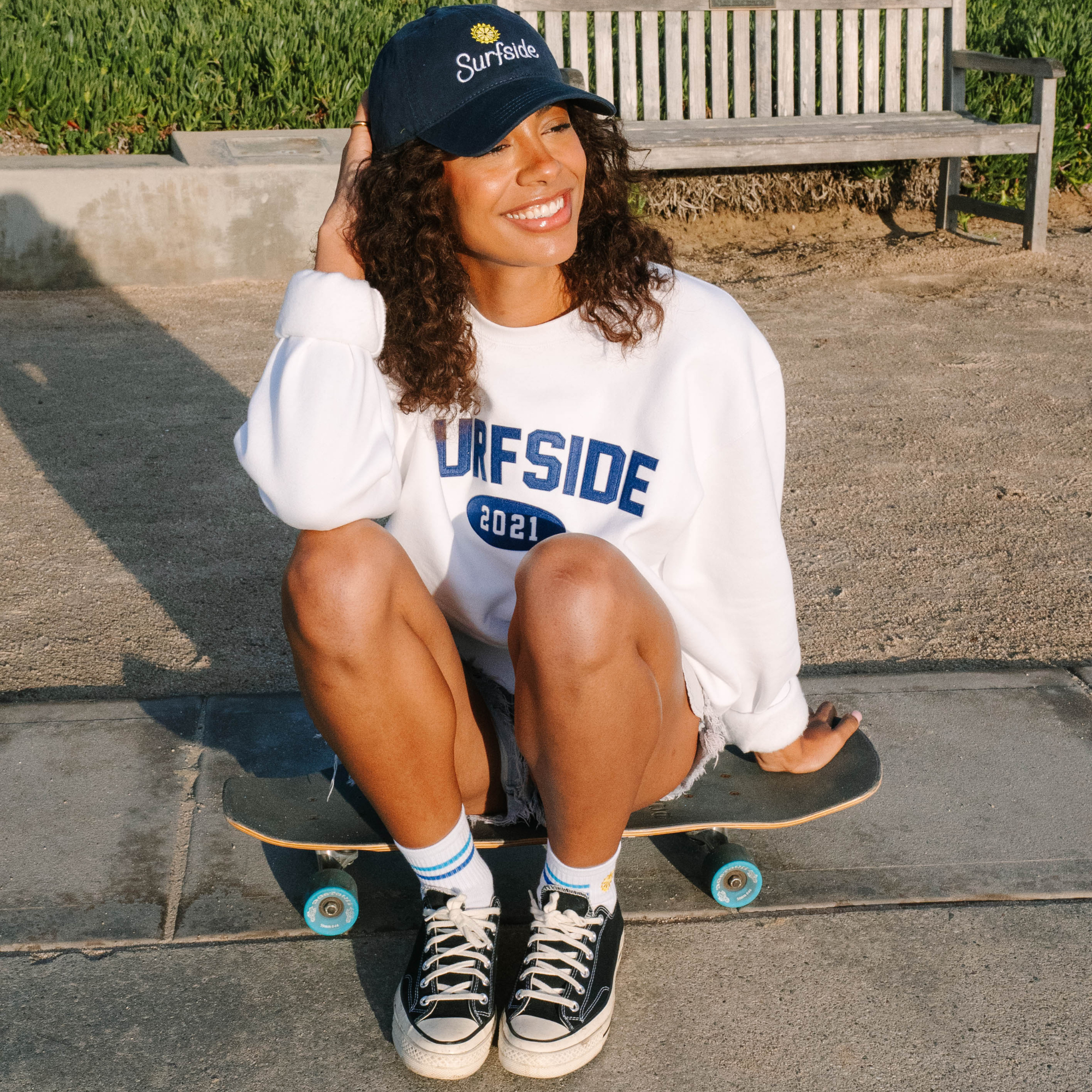 Person sitting on a skateboard wearing a white Surfside 2021 crewneck and a navy Surfside hat in an urban setting.