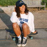 Person sitting on a skateboard wearing a white Surfside 2021 crewneck and a navy Surfside hat in an urban setting.