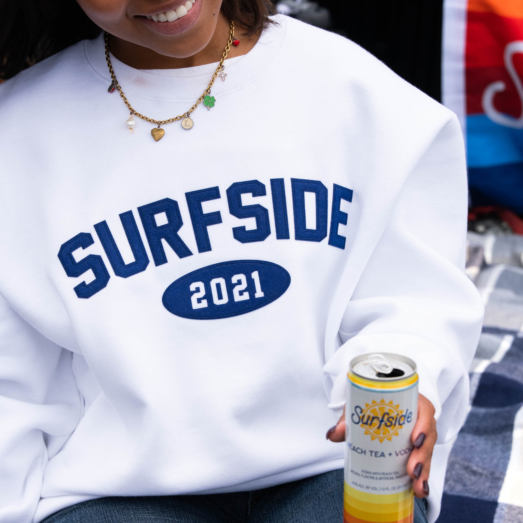 Person wearing a white 'Surfside 2021' crewneck holding a can of 'Surfside Peach Tea and Vodka' product