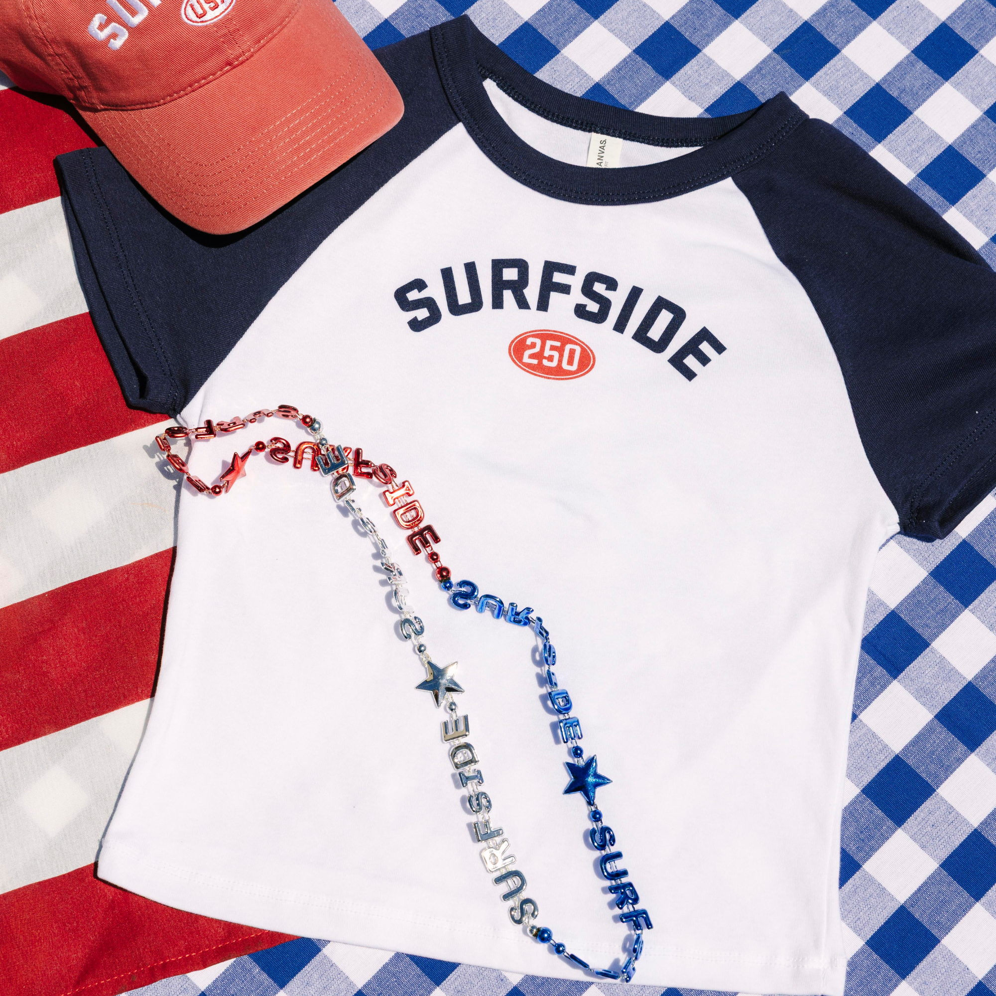 Surfside 250 Baby t-shirt with 'Surfside' print, red cap, and colorful necklace on a checkered background