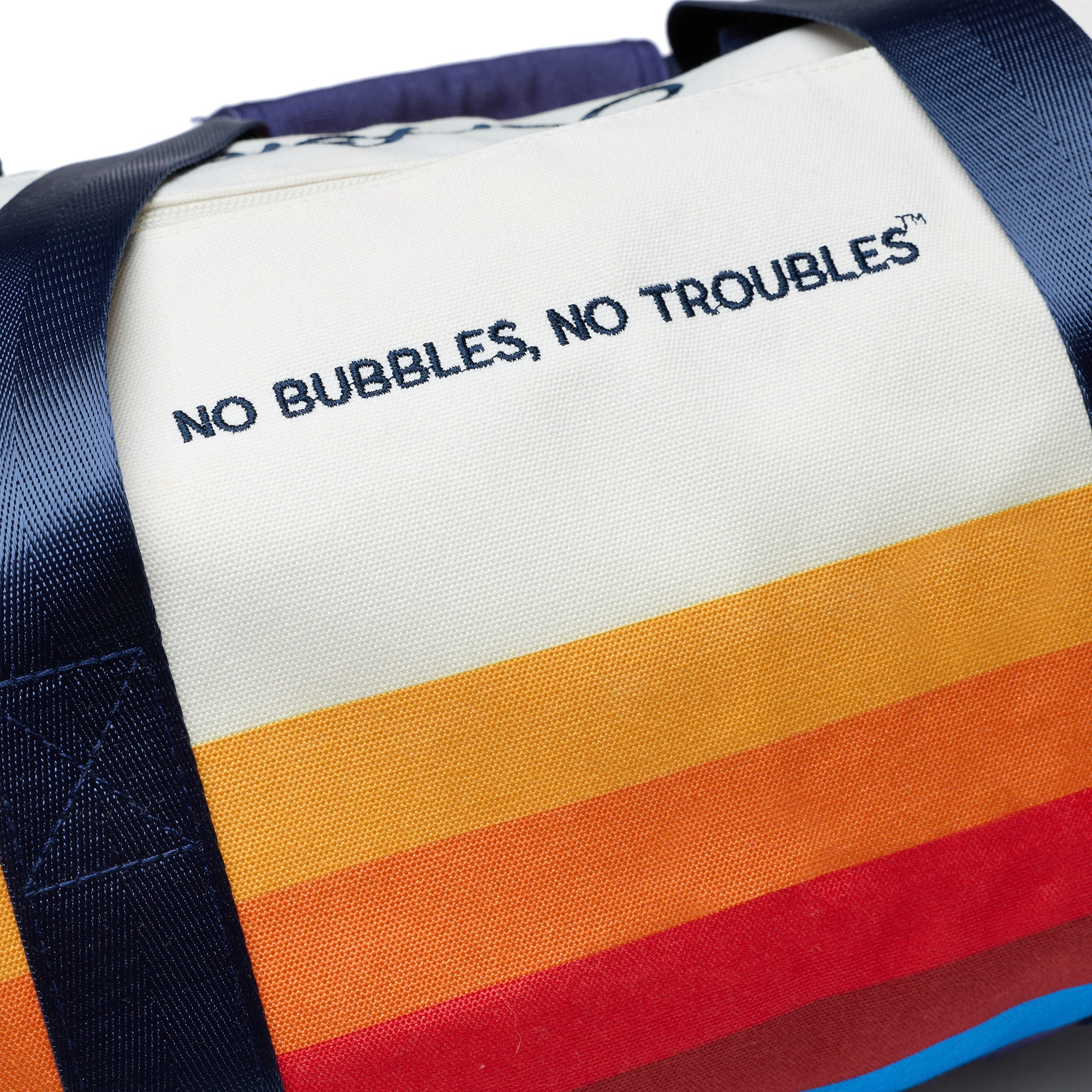 Close-up of a canvas duffle bag with navy blue straps and a cream body, featuring the embroidered text 'NO BUBBLES, NO TROUBLES' and bold horizontal rainbow stripes in orange, yellow, red, and blue.