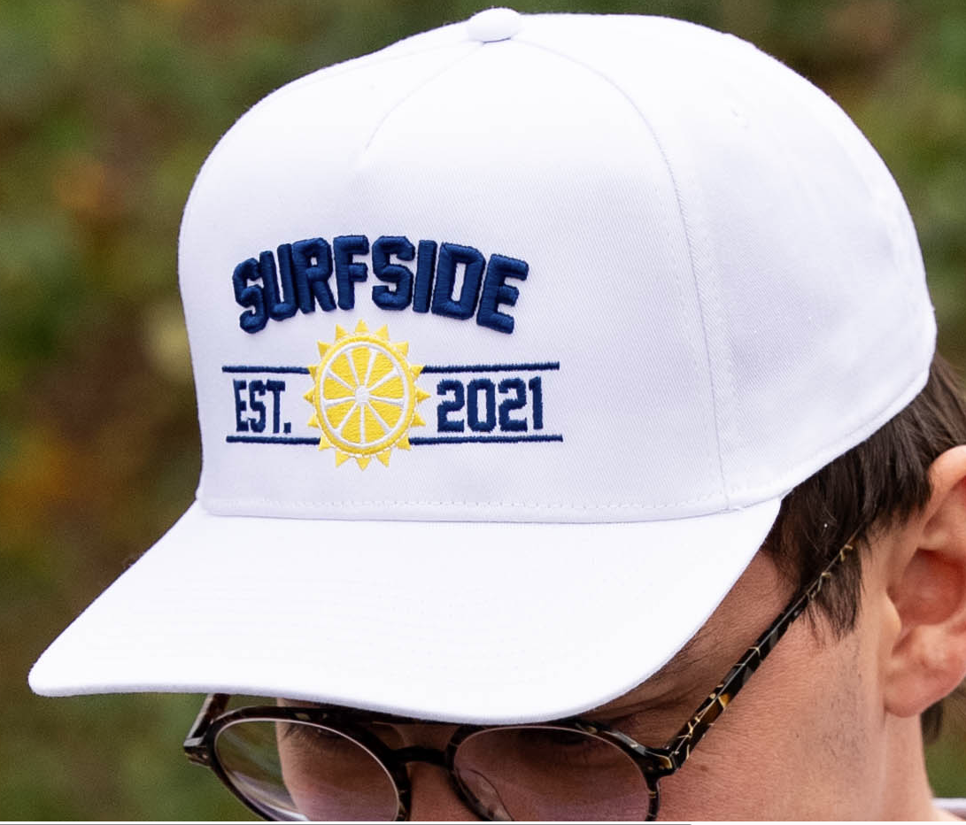 White cap with 'Surfside Est. 2021' text and logo, worn by a person outdoors.