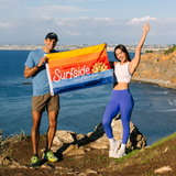 Two people standing on a rocky outcrop by the ocean, holding a 'Surfside' flag.