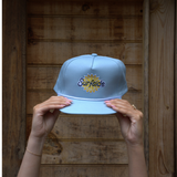 Light blue hat with rope detailing across the brim with 'Surfside' logo held by hands against a wooden background