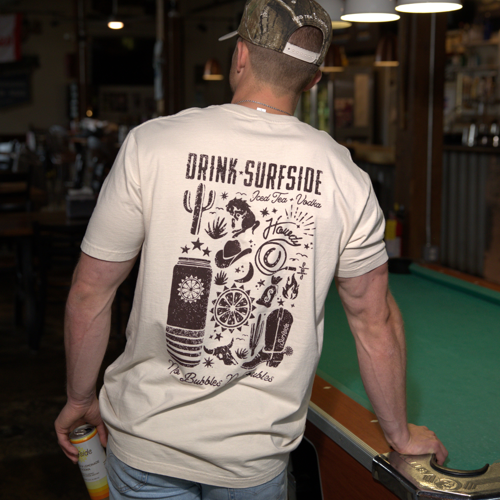 Man wearing a t-shirt with 'Drink Surfside' design and holding a Surfside can in a bar setting