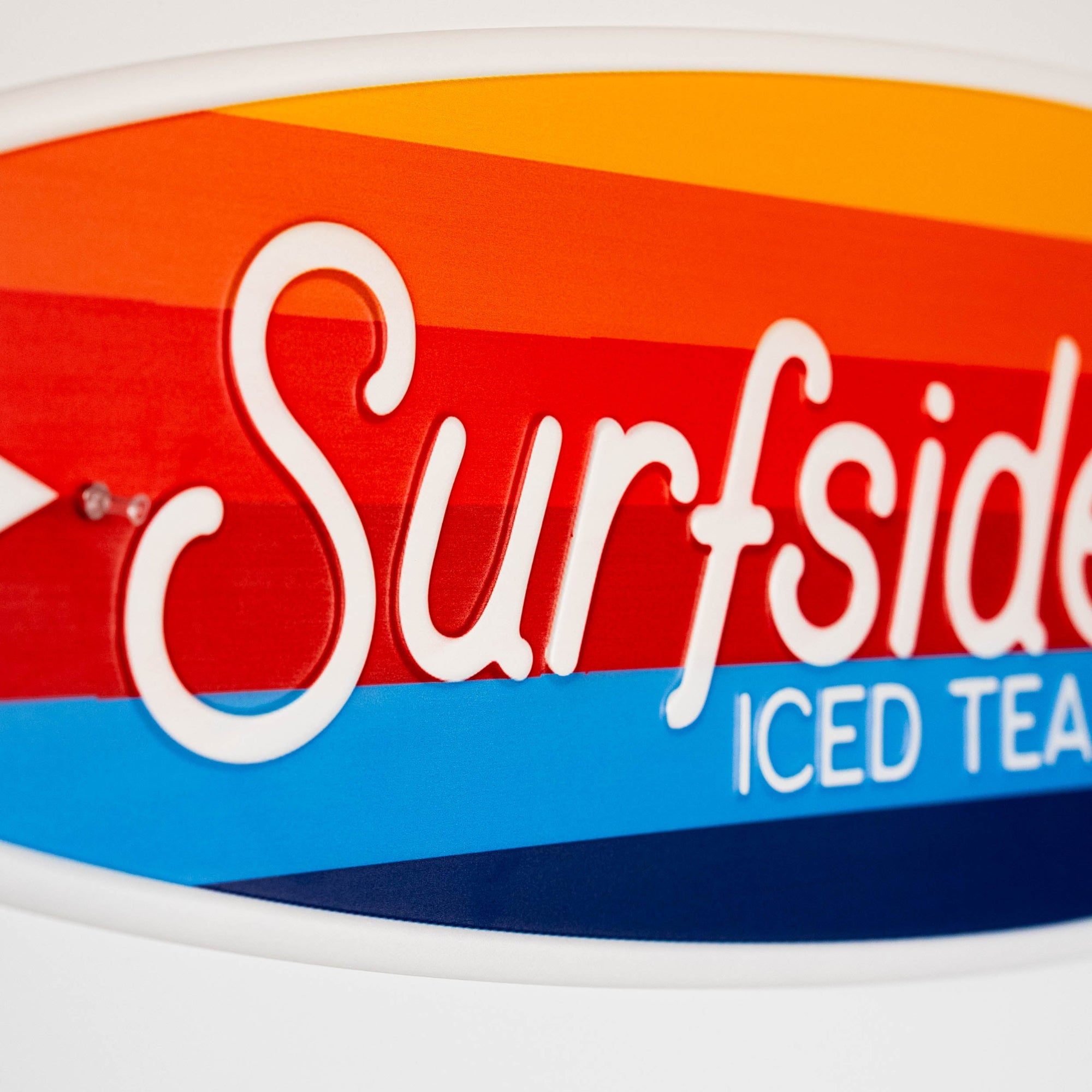 Close up of the Surfside iced tea logo with a colorful design on the surfboard shaped metal sign on a white background