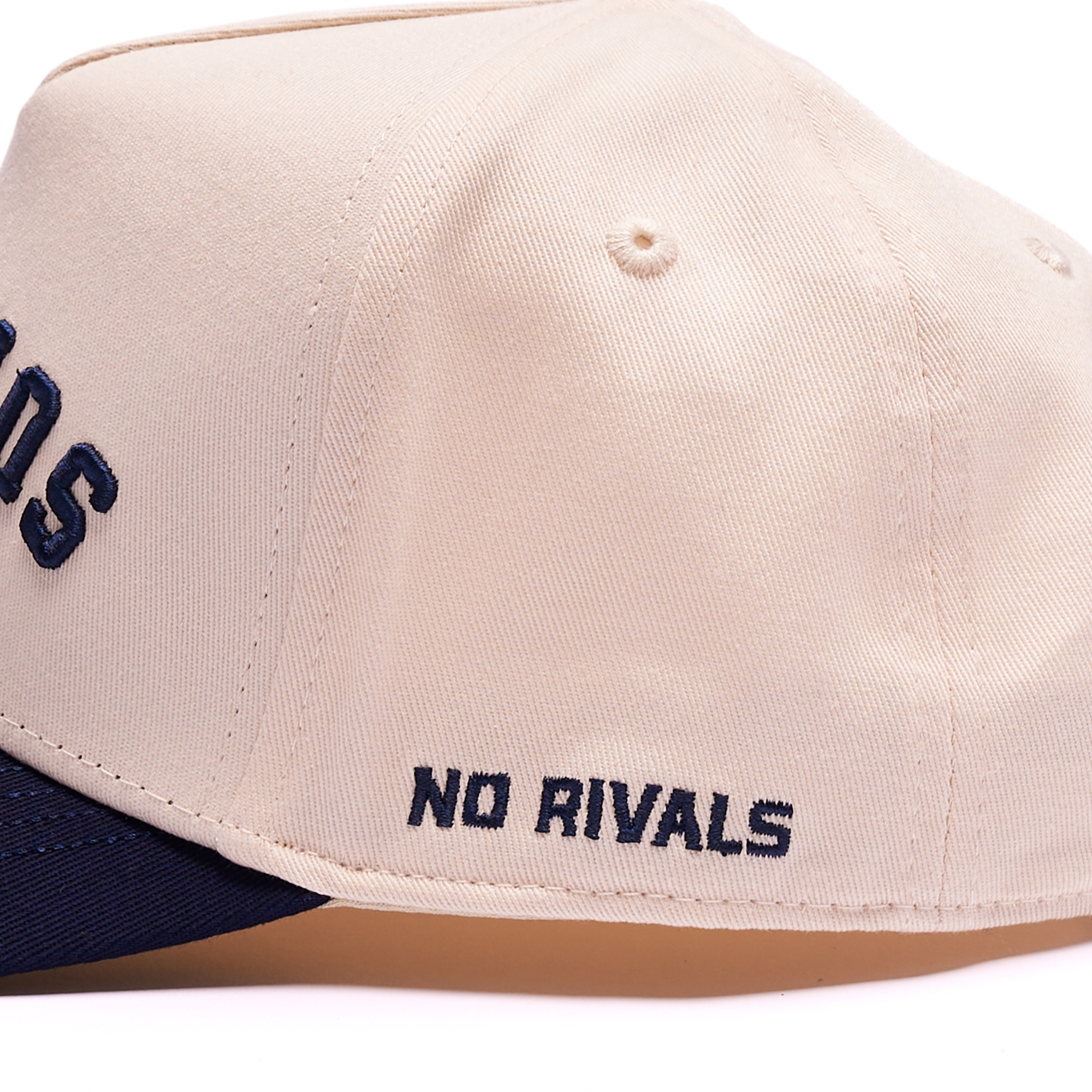 Close-up of a beige snapback cap with navy blue embroidered text reading 'NO RIVALS' on the side and a navy blue visor edge visible at the bottom.
