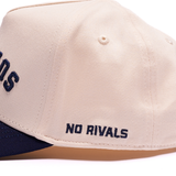 Close-up of a beige snapback cap with navy blue embroidered text reading 'NO RIVALS' on the side and a navy blue visor edge visible at the bottom.