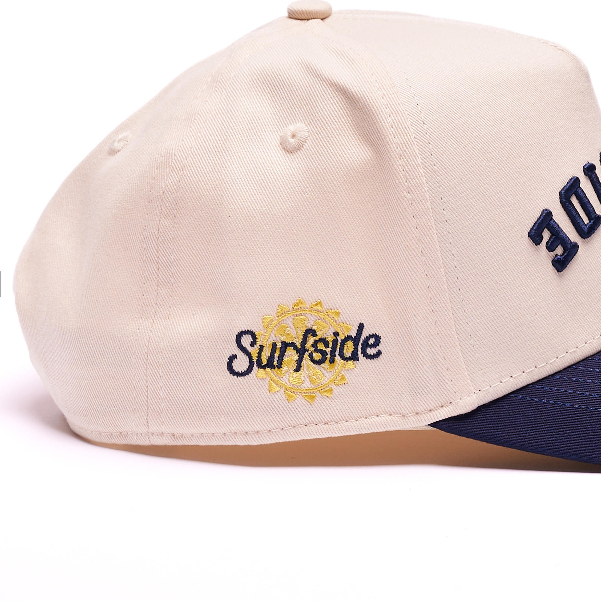 Close-up of a beige baseball cap with navy blue embroidery reading 'Surfside' and a yellow sun emblem on the side, plus partial navy lettering on the front. A navy brim is visible at the bottom.