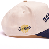 Close-up of a beige baseball cap with navy blue embroidery reading 'Surfside' and a yellow sun emblem on the side, plus partial navy lettering on the front. A navy brim is visible at the bottom.