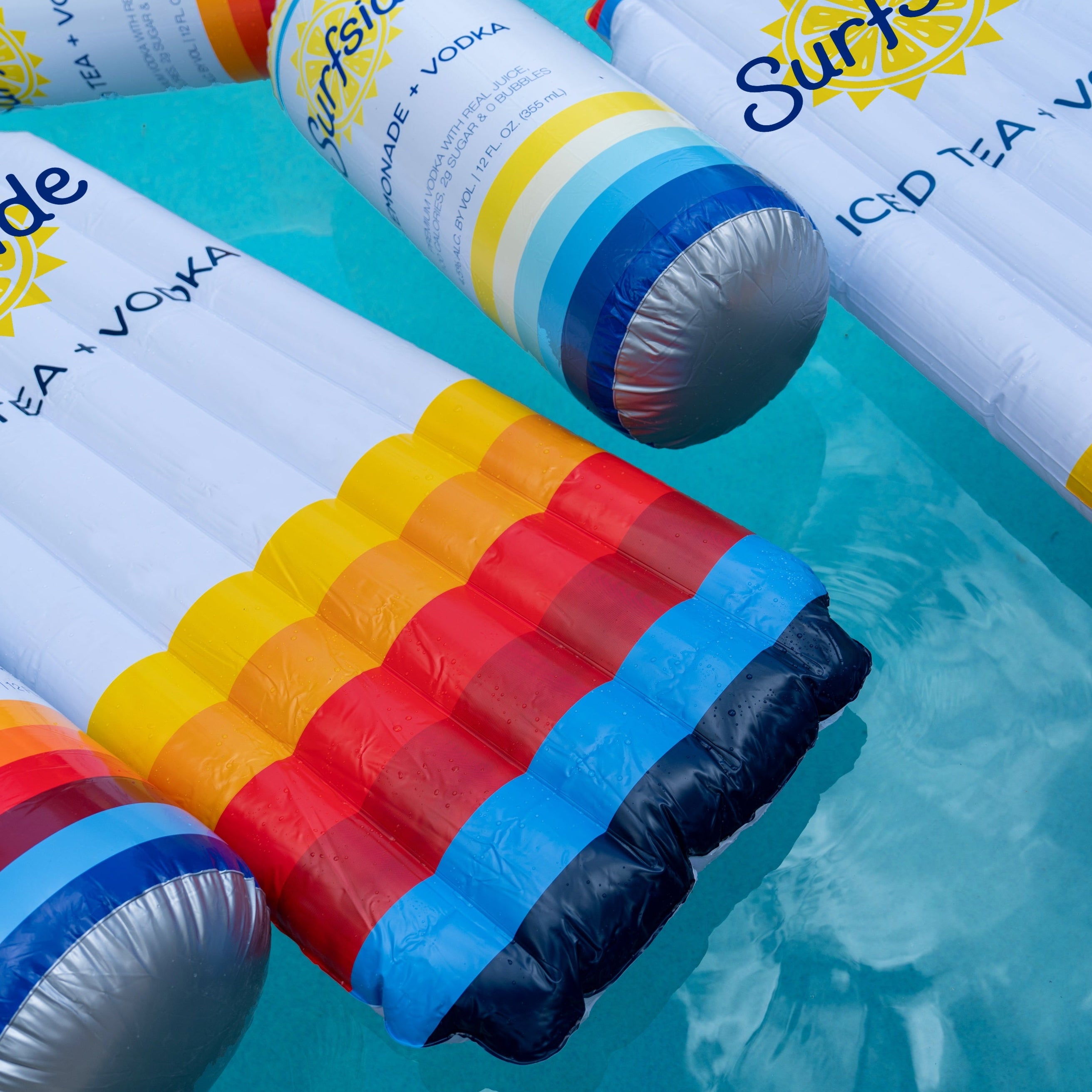 Inflatable pool floats styled like a Surf Tonic vodka can and a striped drink cup, floating on turquoise water. The floats feature white tops with blue and yellow 'Surf Tonic' branding and a bottom section with red, orange, yellow, and blue stripes.