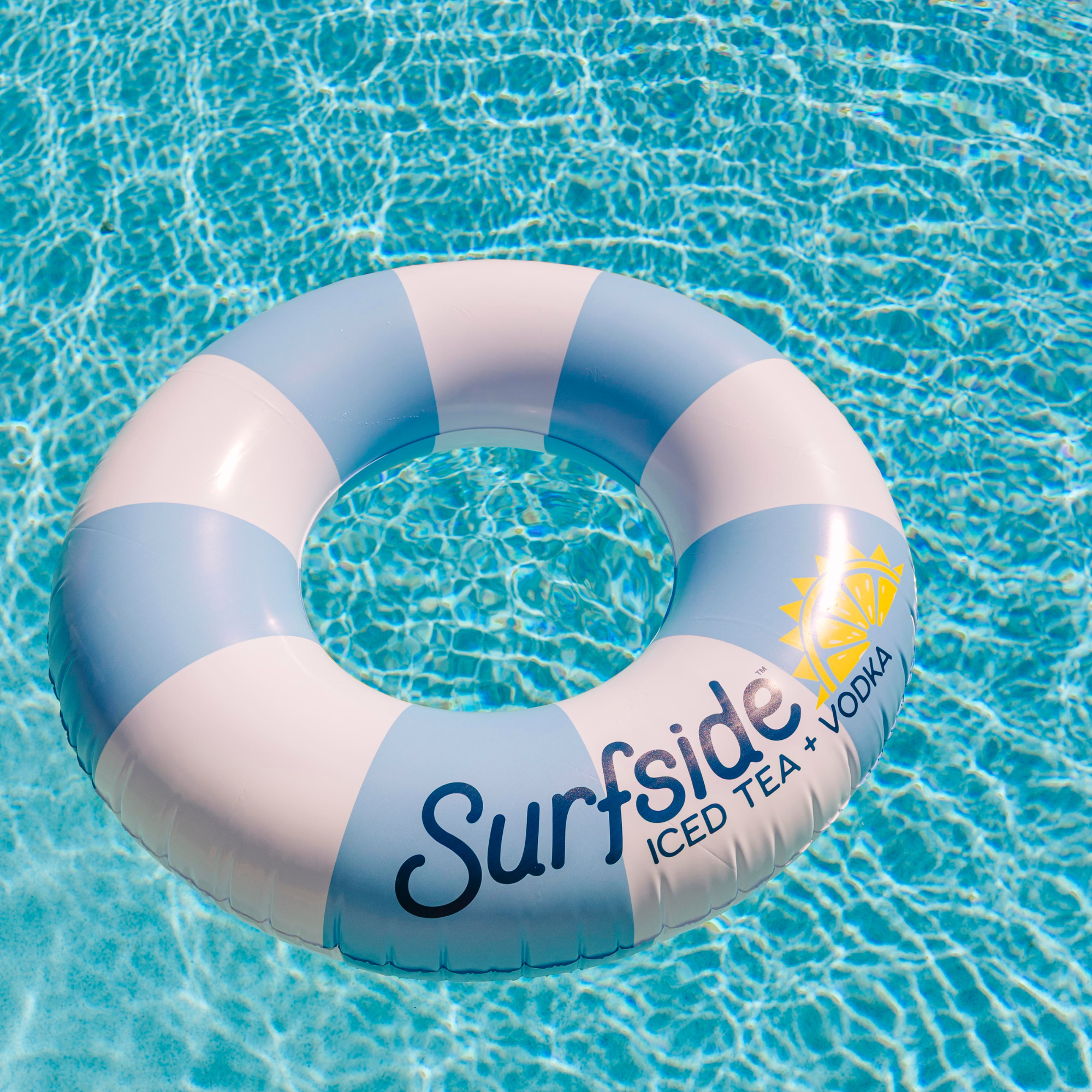 A striped inflatable pool float with light blue and white bands, featuring the text 'Surfside ICED TEA + VODKA' and a yellow sun graphic, floating on clear blue pool water.