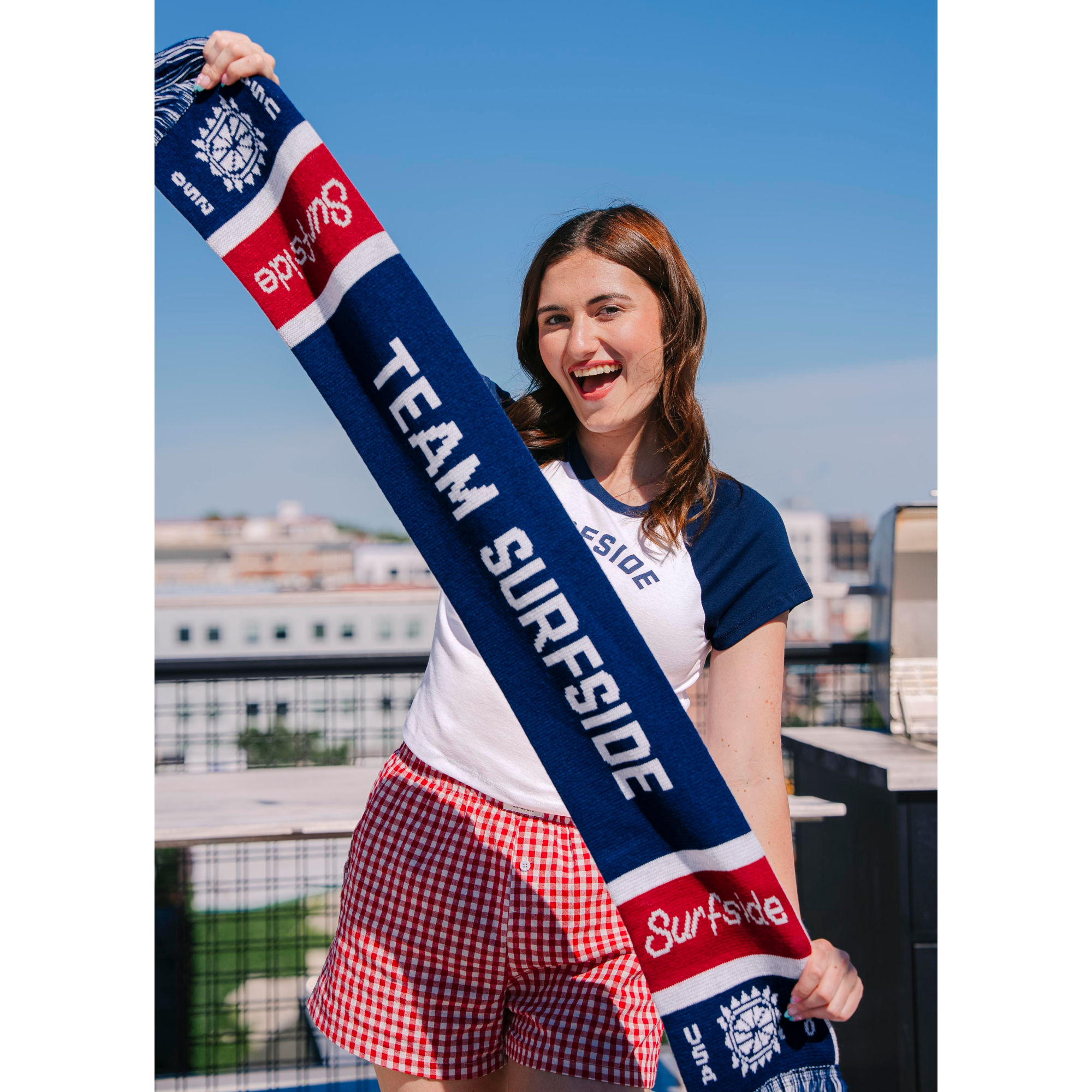 Girl holding Team Surfside USA 250 themed scarf. The Soccer scarf features Team Surfside in the middle, the surfside logo and the sun logo with fringe. The soccer scarf is red, white and blue.